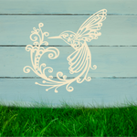 Delicate metal wall art featuring a hummingbird with intricately detailed wings and tail, surrounded by swirling vine patterns, finished in Canola Cream powder coating, mounted on a rustic white wooden backdrop, perfect for adding a touch of nature to an outdoor space.